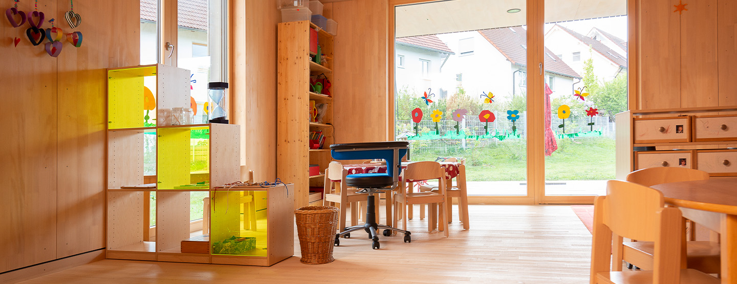 Innenraum einer Kita in moderner Holzbauweise mit Holztischen, Holzstühlen und Regalen – nachhaltiger Holzbau für Kommunen, Kitas und Schulen im Neubau.