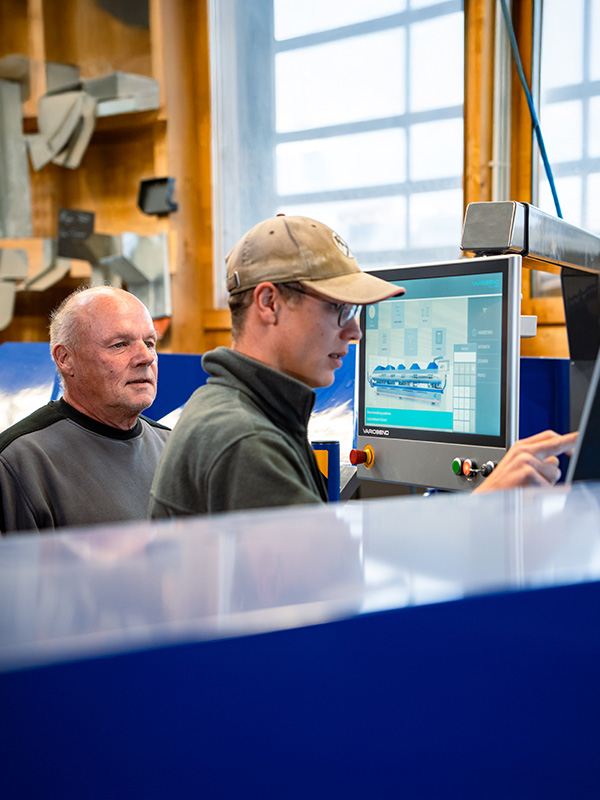 Älterer Ettwein‑Mitarbeiter erklärt Bedienung am Maschinen‑Display einem Auszubildenden — Ausbildung, Schreiner, Zimmerer, Dachdecker, Handwerk.