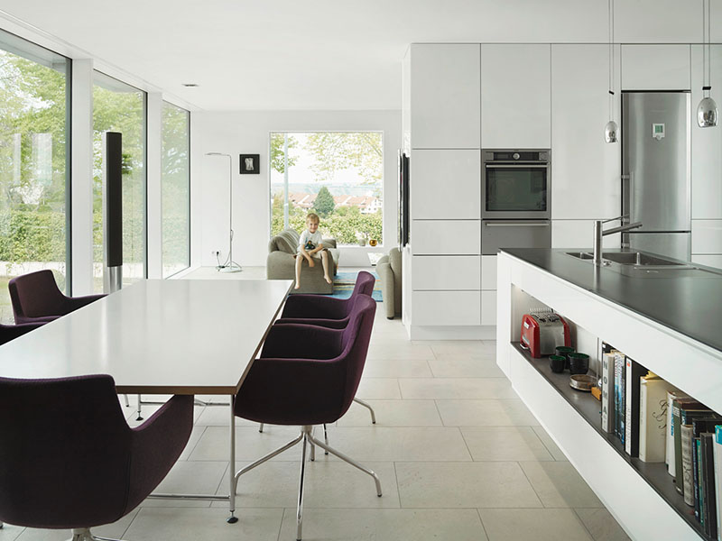 Weiße Holzküche mit grauer Arbeitsfläche, weißen Schränken, weißem Tisch und violetten Stühlen. Im Hintergrund Wohnzimmer mit großem Fenster und Junge auf Sofa.