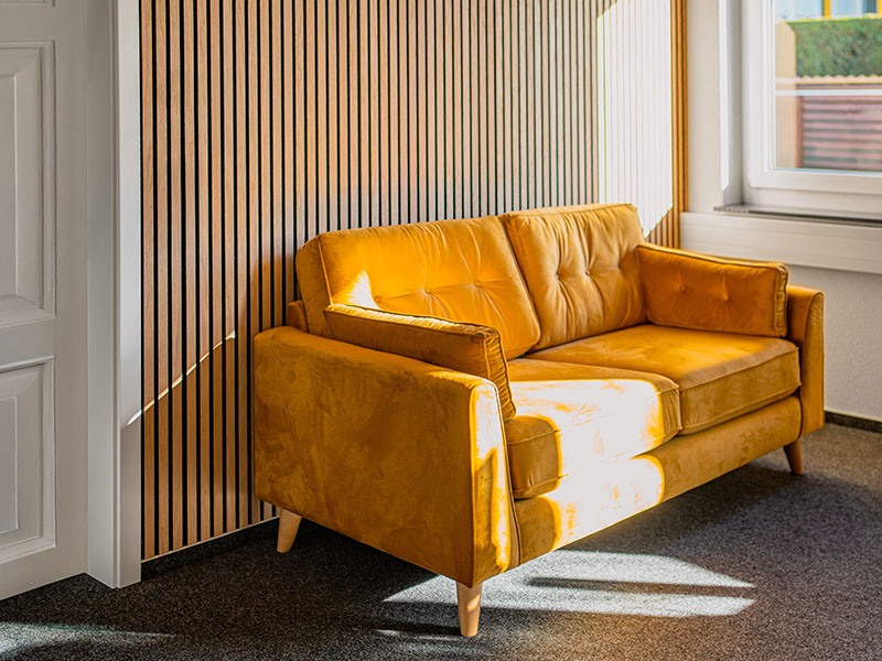 Gelbes Sofa vor heller Akustikpaneele aus Holz mit einfallendem Sonnenlicht — Akustikpaneele von Lignotrend, Wandpaneele und Deckenpaneele.