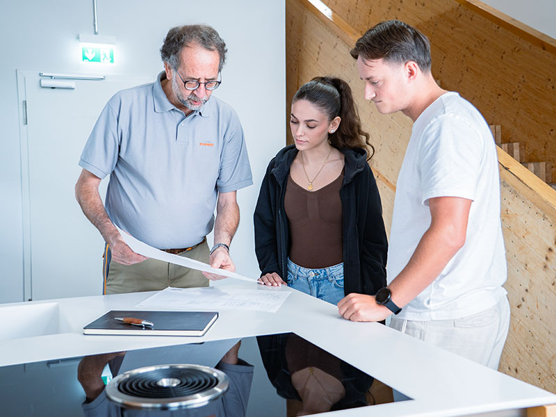 Ettwein-Mitarbeiter berät junges Paar zur neuen Holzküche an Muster-Arbeitsplatte mit Herd, Buch, Stift und Küchenplan.