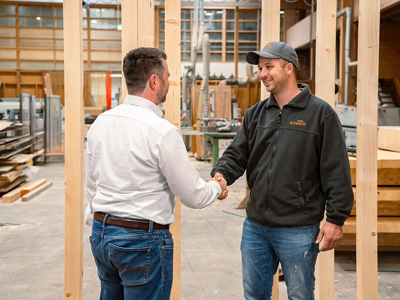 Ettwein-Mitarbeiter schüttelt einem Kunden in der Fertigungshalle die Hand – Vertrauen und Partnerschaft im Holzbau für Kommunen, Kitas, Schulen und Neubauten.