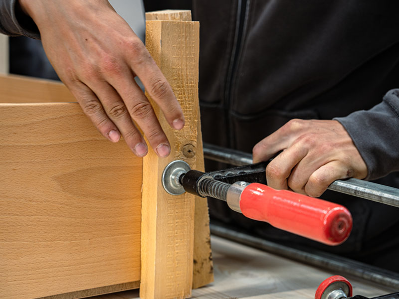 Nahaufnahme von Händen an einem Holzstück mit eingesetzter Schraubzwinge — Ausbildung, Schreiner, Zimmerer, Dachdecker, Handwerk.