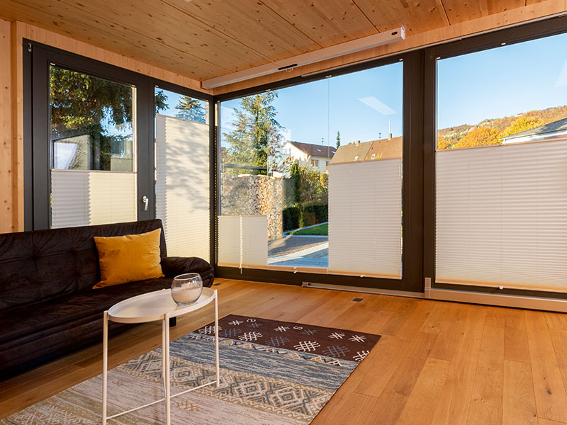 Innenansicht eines Ettwein Mobile Room mit Holzfußboden, Holzdecke, Sofa und großer Fensterfront – nachhaltiger Holzbau aus der Zimmerei und Schreinerei Ettwein.
