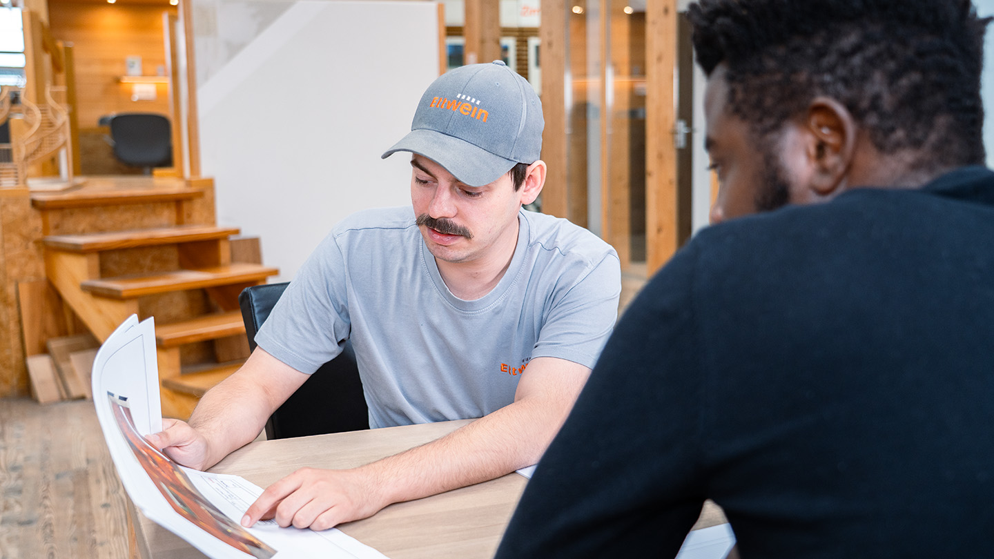 Ettwein-Mitarbeiter berät Kunden im Firmengebäude und zeigt eine Visualisierung des Projekts auf Papier – persönliche Beratung rund um das Holzhaus, Traumhaus, Neubau und Umbau.