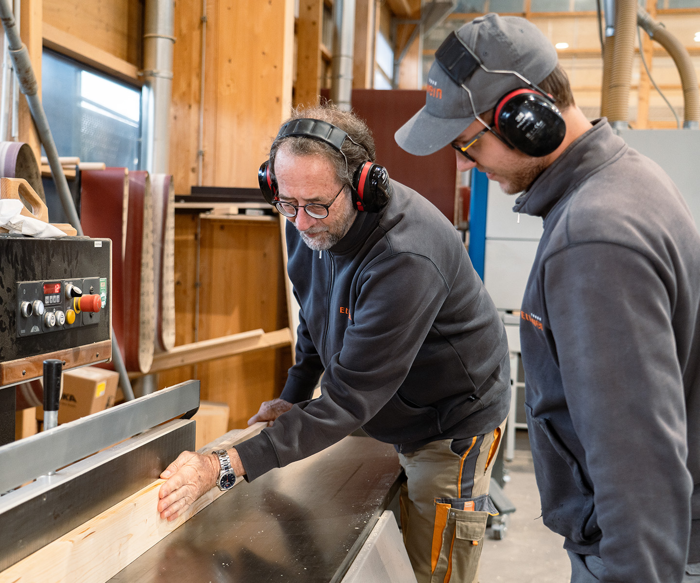 Älterer Ettwein‑Mitarbeiter zeigt einem jüngeren an einer Maschine etwas; beide tragen Gehörschutz — Ausbildung, Schreiner, Zimmerer, Dachdecker, Handwerk.